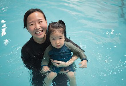 A woman holds a child in shallow water, both smiling and enjoying a swim at the pool.