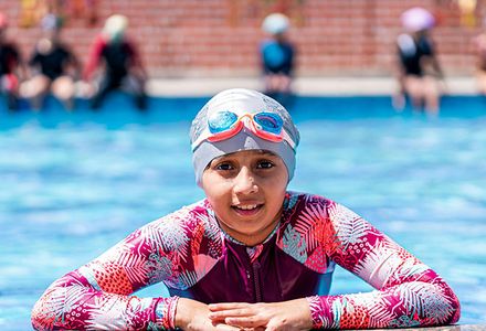 A young girl wearing bathers, a swimming cap and goggles looks into the camera from a swimming pool.
