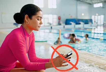 A woman plays on her phone at the swimming pool whilst children play in the background. A red cross over her indicates she should be focusing her attention on the children she is supervising.